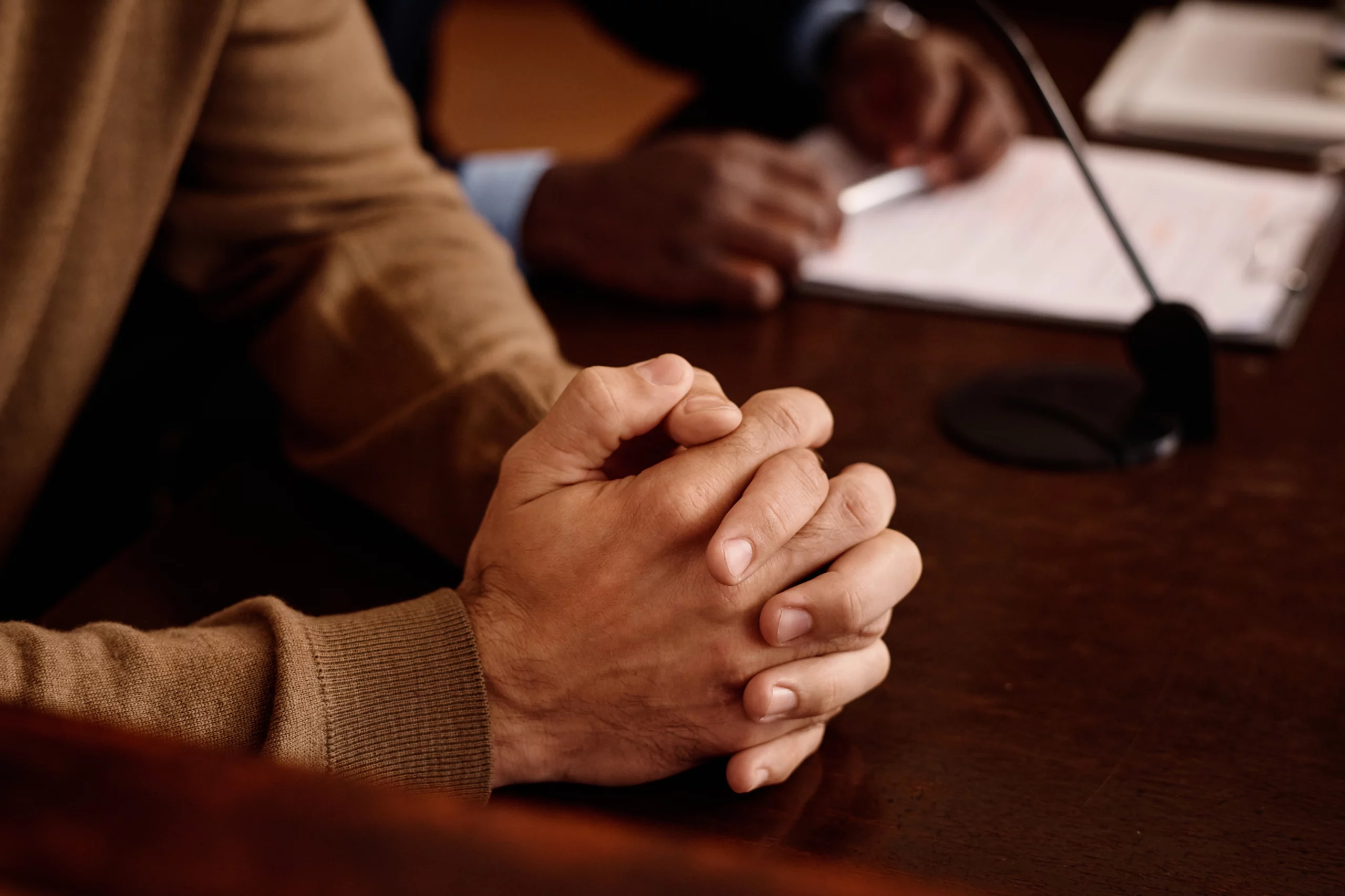 Two people seated at a table; one with clasped hands, the other writing on paper, with a microphone indicating a formal interview or testimony setting