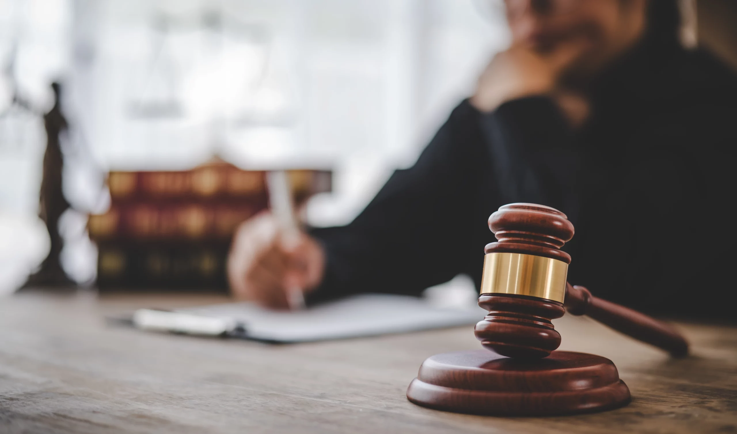 Gavel on desk with blurred person writing behind law books and justice scales.