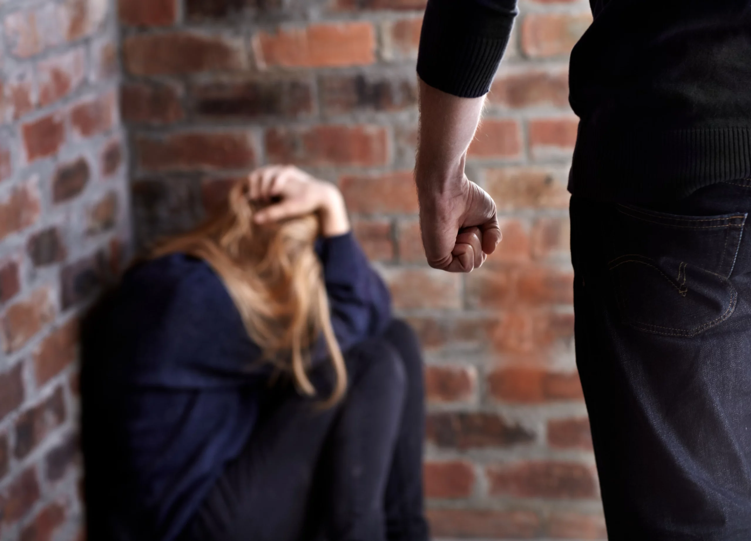 Frightened person cowers in a corner, shielding their head while a clenched fist looms threateningly in the foreground—evoking tension and fear.