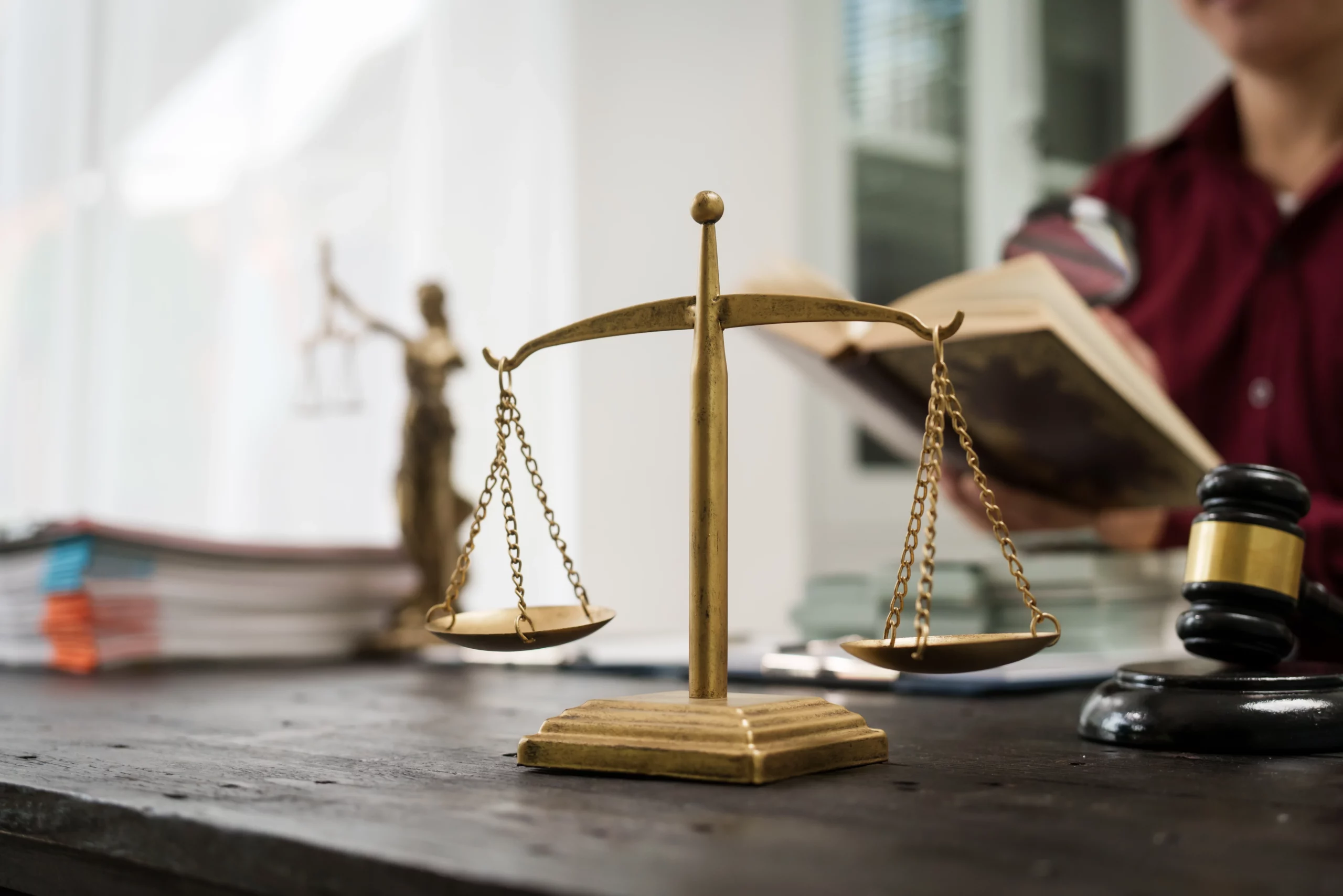 Close-up of golden balance scale, gavel and open legal book on lawyer’s desk, symbolizing justice and legal challenge