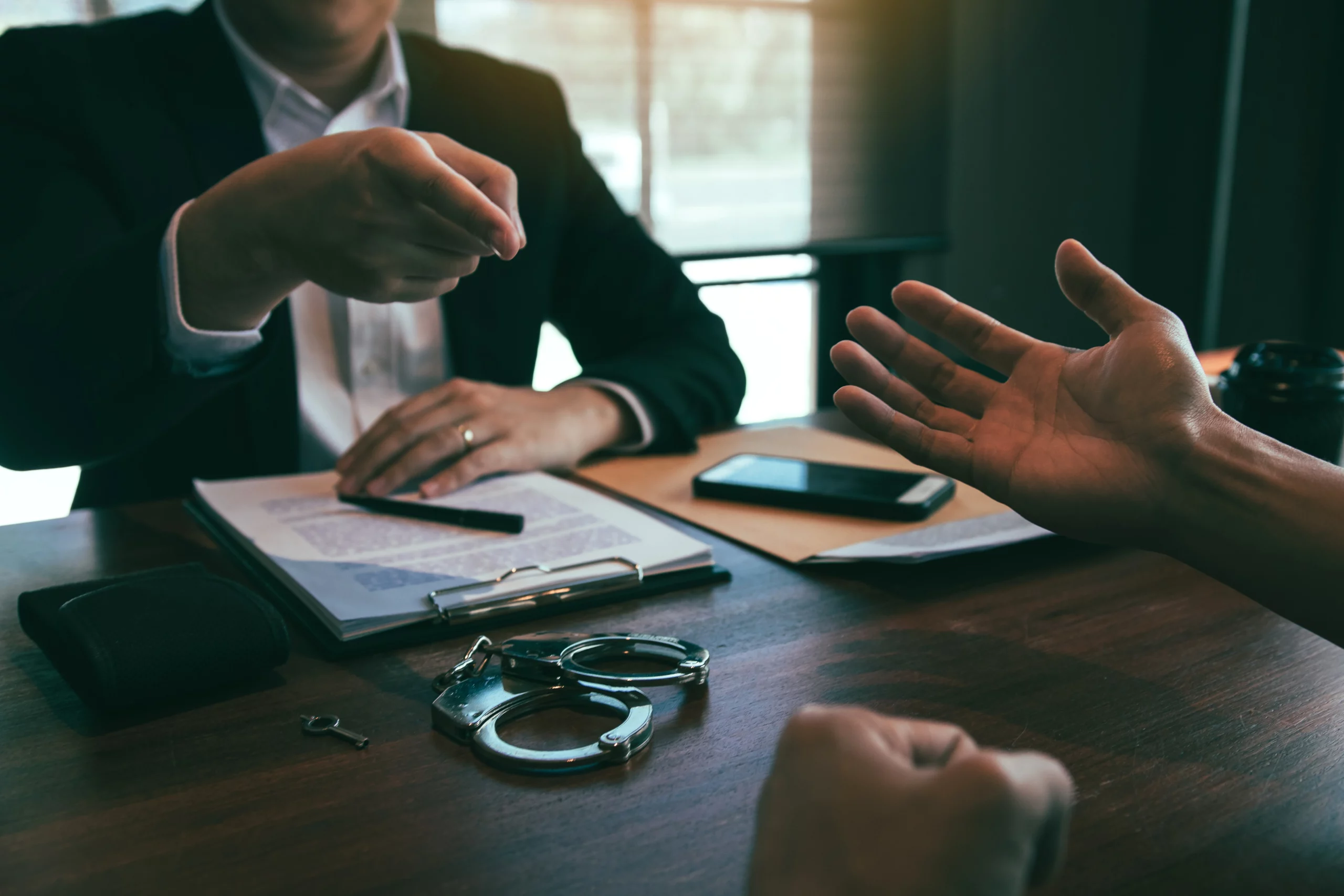 Person reviewing legal document with pen while another gestures; handcuffs and key on table symbolize criminal charge discussion