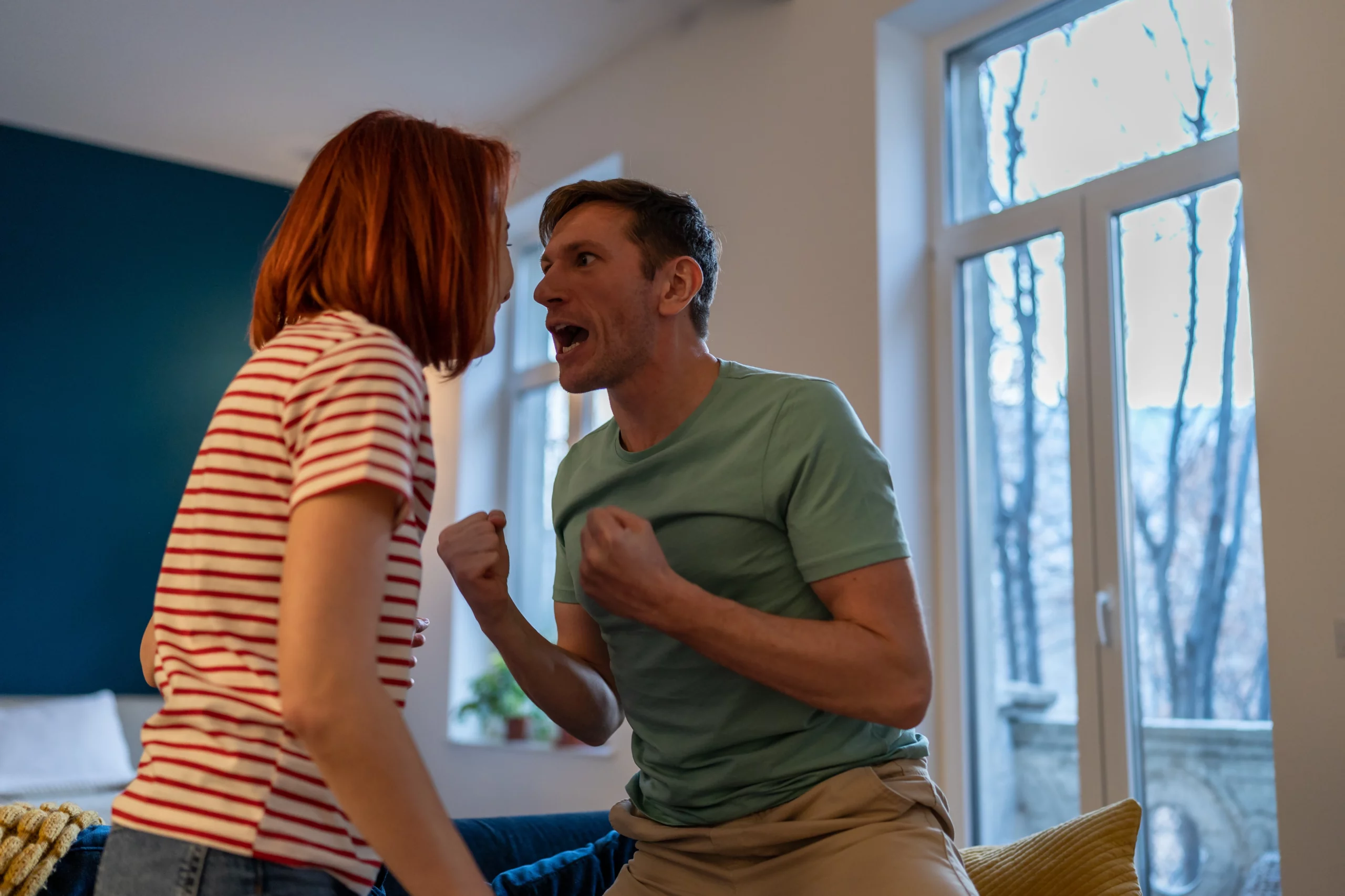 Man and woman in heated argument at home, man raising fists in anger, capturing conflict and escalation