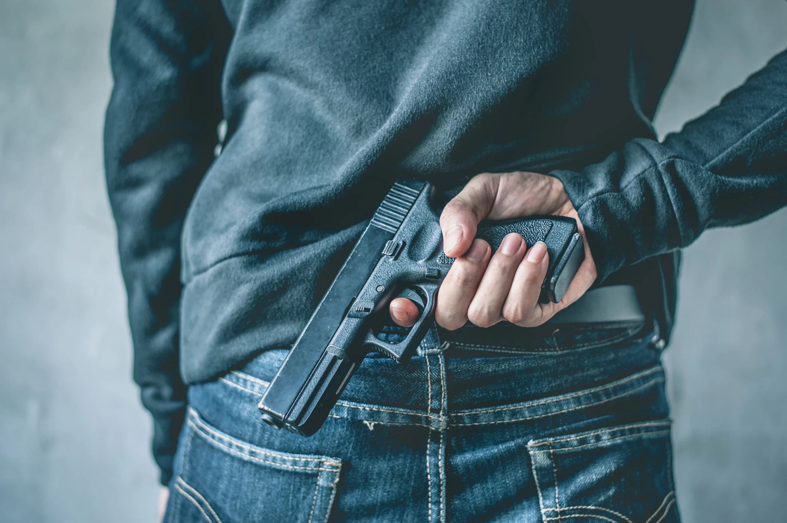 Person in dark sweater and jeans holding a handgun behind their back against light gray wall, illustrating firearms offenses charged under Indiana and federal law.